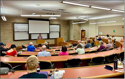 2024-10-08_Universe-and-Human-Stupidity_400px.png speaker at podium addressing participants in classroom
