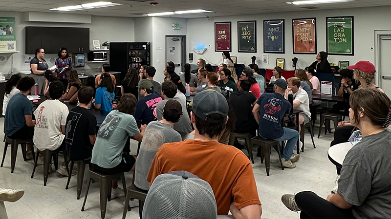 Inset_2025-Day-of-Service_800x450.png students seated around table in conference room in front of two speakers, standing